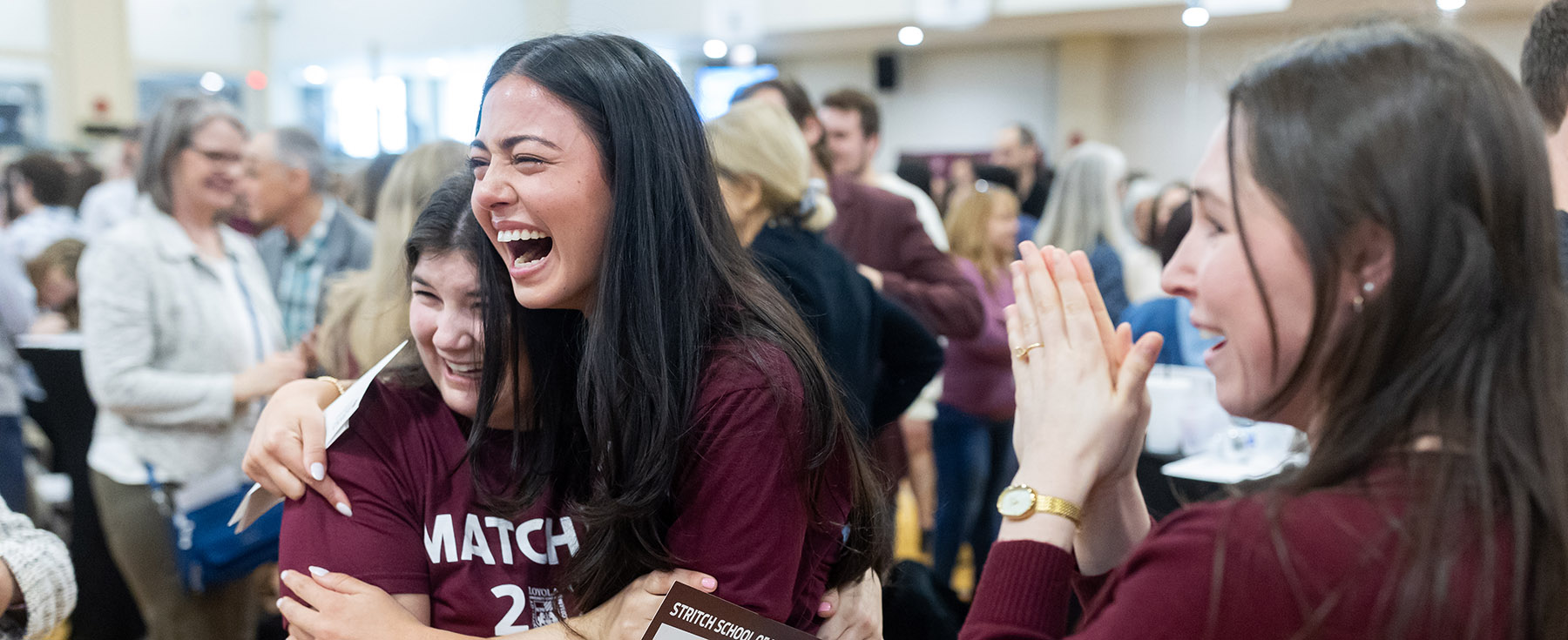 Two students wearing maroon t-shirts embrace while a student claps next to them